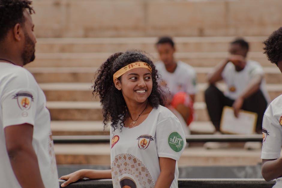 A group of young adults smiling and standing outdoors, wearing white shirts with logo designs.