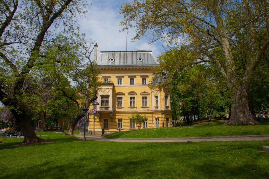 Yellow historic building surrounded by lush park and trees under a clear sky.