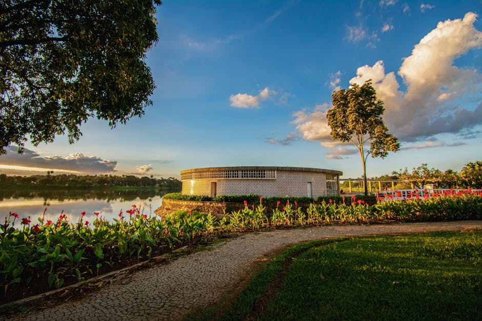 Beautiful landscape with a circular building by a lake, surrounded by lush greenery and colorful flowers at sunset.